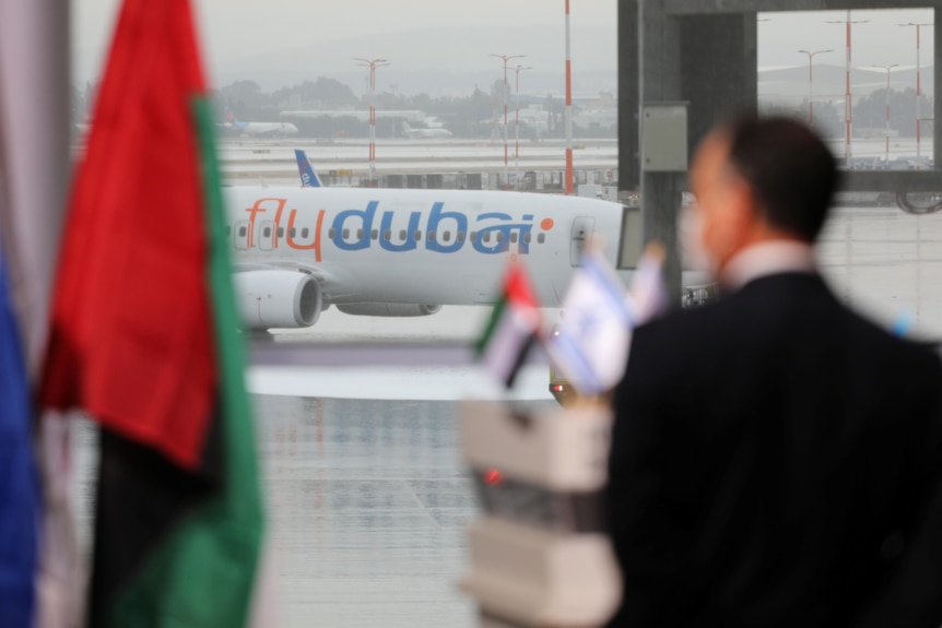 A FlyDubai aircraft is seen through a window, with a UAE flag and a man in the foreground.