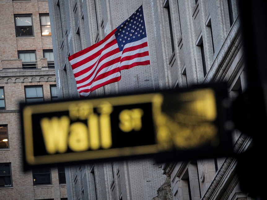 The US flag is flying on the side of a building with a blurred street sign for Wall St in the foreground.